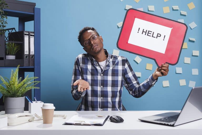 A person in a checkered shirt sits at a desk, holding a red sign with the word "HELP" in a speech bubble. Post-it notes cover the blue wall behind them, perhaps reminders for an upcoming meeting with their financial planner.