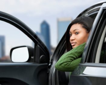 A person wearing a green sweater sits in a car with the door open, looking out. Urban buildings are visible in the background, evoking a nostalgic Gen X vibe.