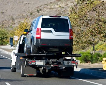 A silver SUV, likely a car repo, is being towed on a flatbed tow truck along a road, surrounded by dry vegetation and hills in the background.