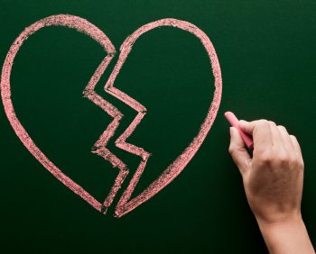 A hand is drawing a pink broken heart on a green chalkboard with chalk, symbolizing the pain of divorce.