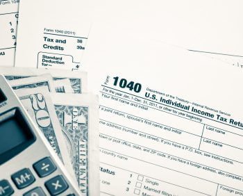 A calculator, U.S. Individual Income Tax Return forms (1040), and taxable dollar bills are arranged on a table.