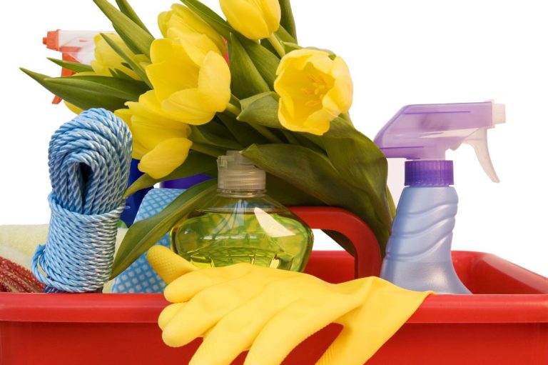 A red basket filled with cleaning supplies, perfect for a spring clean, includes a spray bottle, gloves, detergent, a coiled rope, and a bunch of yellow tulips.