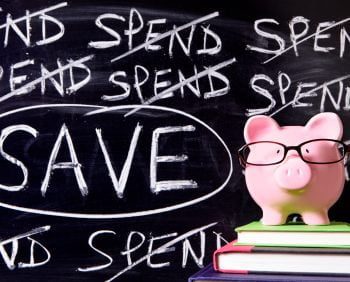 A pink piggy bank with glasses sits on a stack of books in front of a chalkboard with the word "SPEND" crossed out multiple times and "SAVE" circled prominently, offering a perfect visual for summer financial wisdom.