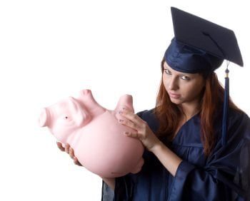 A graduate in a navy blue cap and gown holds an upside-down pink piggy bank, looking concerned about student loans.