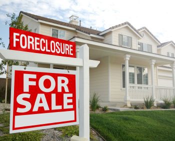 A two-story house with a front yard showcases a "Foreclosure" and "For Sale" sign planted in the foreground.