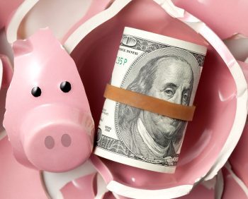 A roll of U.S. hundred dollar bills secured with a rubber band lies among pieces of a broken pink piggy bank, symbolizing shattered dreams and lost retirement money.