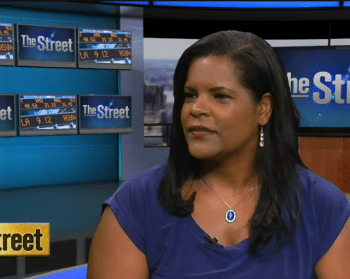 A woman in a purple top appears in a professional studio interview with "The Street" logo visible in the background, as screens display stock market information and budget reports.