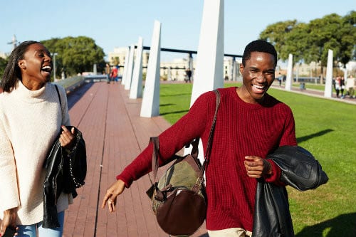 Two people stroll along a brick path in the park, laughing and carrying bags, discussing their plans with a financial planner.