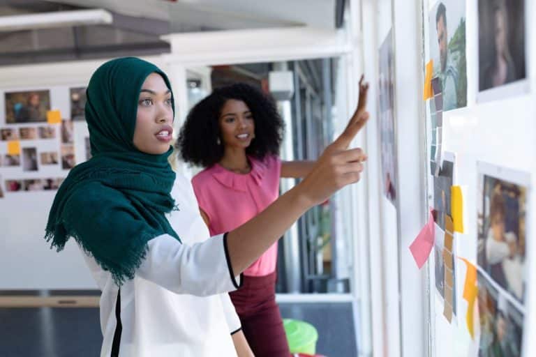 Two women collaborate in an office, looking at photos and notes on a wall. One, a financial planner, points to a specific spot on the board as they discuss budget strategies.