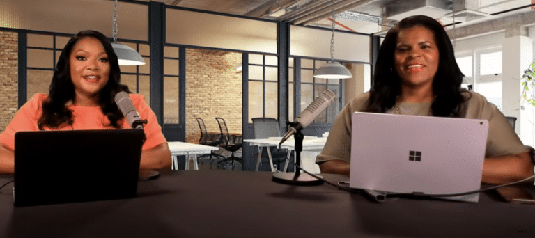 Two women sit at a table with laptops and microphones, discussing budget strategies in a modern office setting with brick walls and pendant lights.