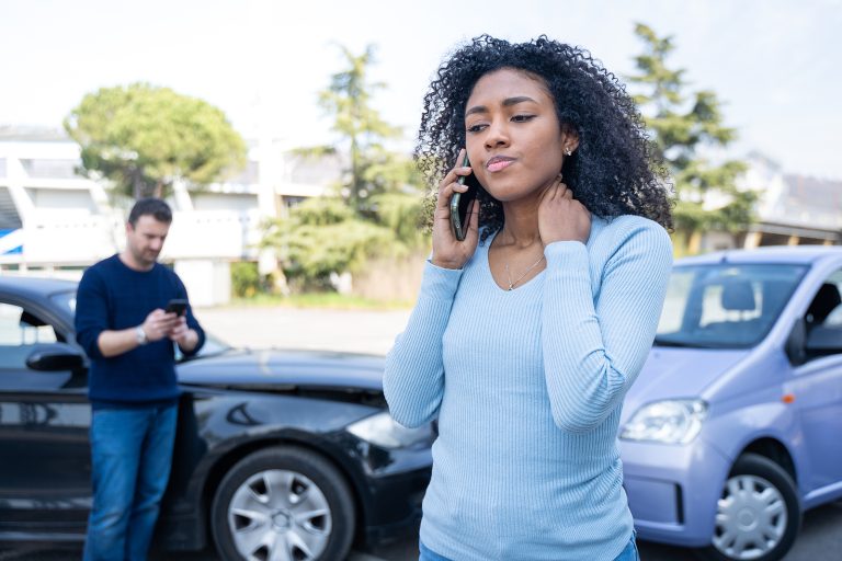 Two cars are involved in a minor accident. A woman, possibly calling her financial planner, stands in the foreground on the phone, while a man checks his phone near the damaged vehicles.