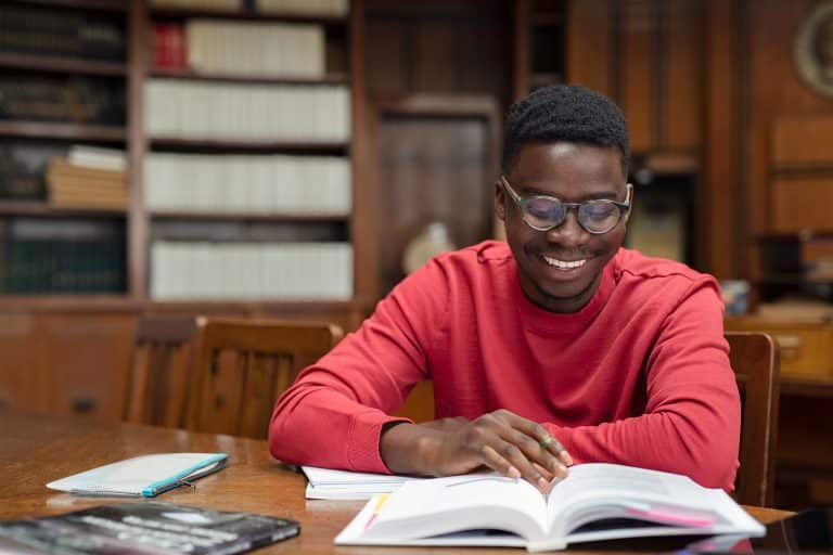 A person wearing glasses and a red sweater is sitting at a wooden table, reading a book on financial planning with papers nearby. Shelves filled with books are in the background, creating an ideal setting for budget insights.