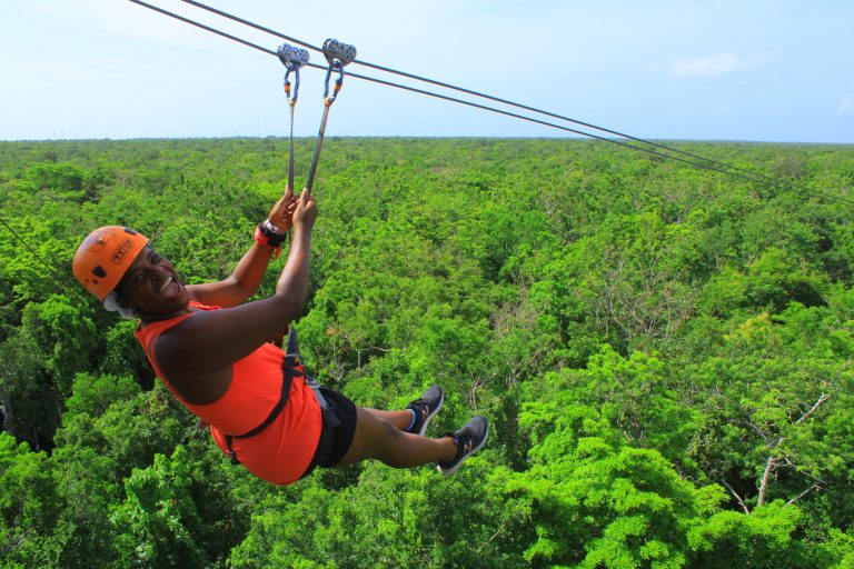 A person ziplining over a lush green forest, smiling broadly in an orange helmet and shirt, savors the thrill of adventure without worrying about budget constraints or credit concerns.