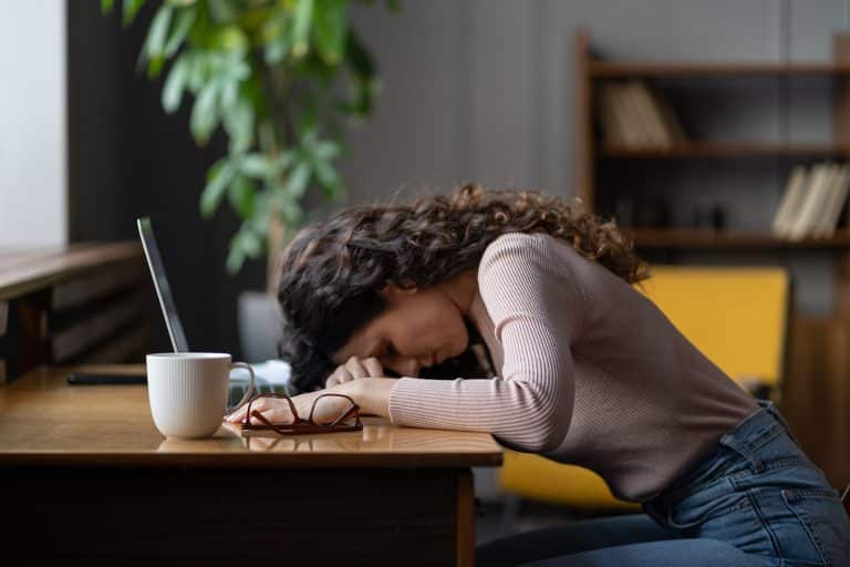 A woman rests her head on a desk, with a laptop, mug, and glasses nearby—surrounded by bookshelves and a plant. As if taking a break from meticulous budgeting or tackling the complexities of credit management, she finds solace in this moment of pause.