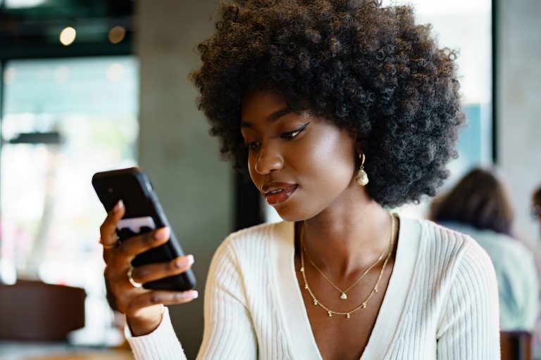 A woman with curly hair, likely a financial planner, is sitting indoors, intently looking at her smartphone. She is wearing a white top and gold jewelry, possibly reviewing budgets and credit options for her clients.