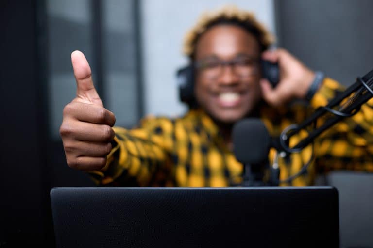 A person in a yellow plaid shirt and headphones gives a thumbs-up while sitting at a desk with a microphone, laptop, and financial planner.