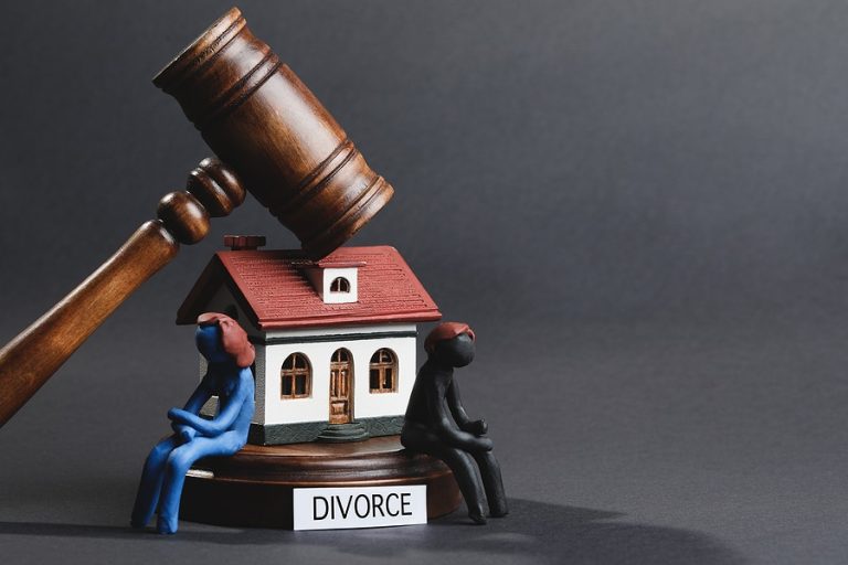 A wooden gavel hovers over a model house with two figurines sitting back-to-back, symbolizing the division of assets. A label below reads "Divorce," highlighting the importance of consulting a financial planner to manage credit and budget effectively during such transitions.