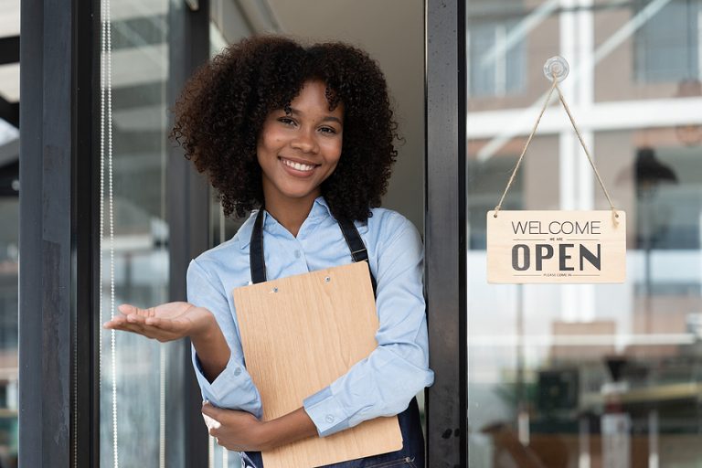 A smiling financial planner with curly hair holds a clipboard, standing next to a "Welcome, Open" sign at the store entrance.