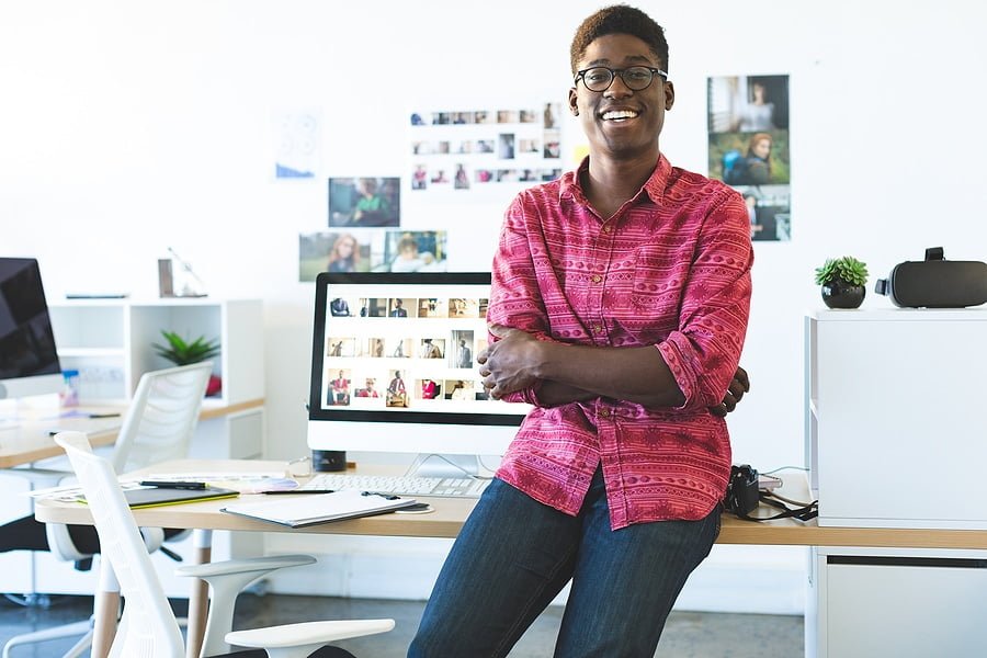 Portrait of young African-American graphic designer looking and smiling at camera while sitting at desk in office