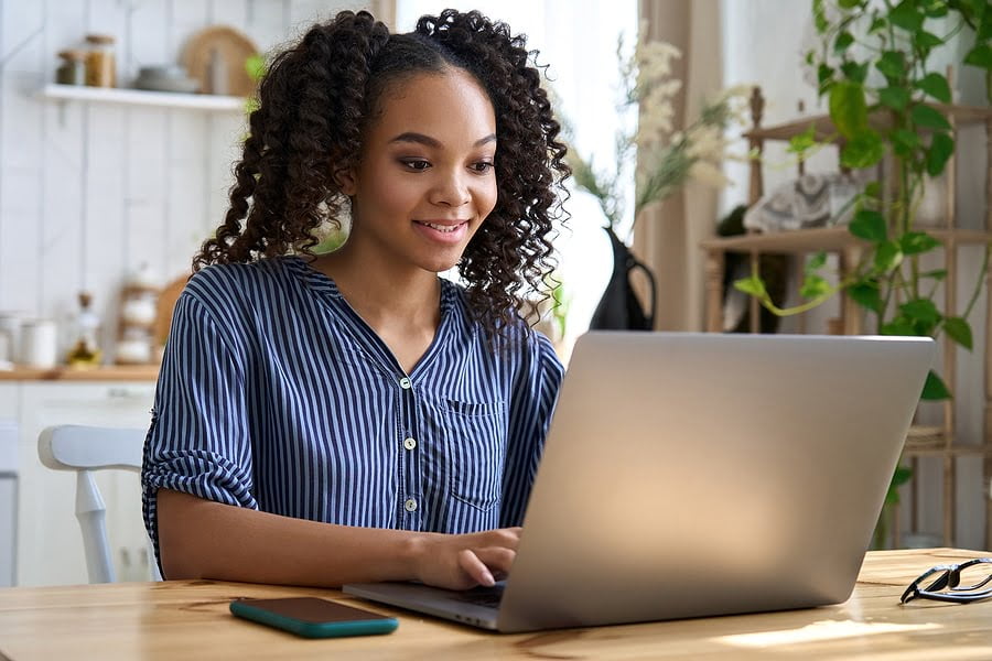 Smiling teenage girl college student using laptop computer sitting at table at home learning online or browsing internet typing on computer, studying