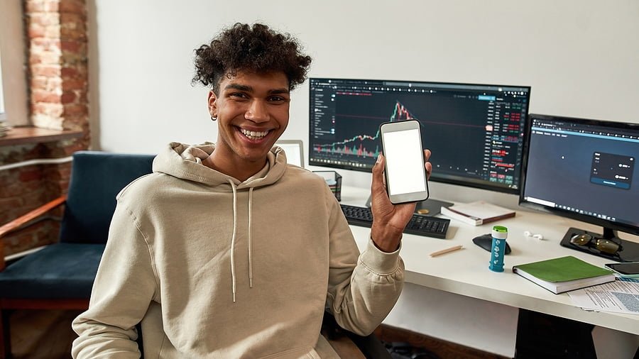 Web wide portrait of happy African American man investor recommend financial app on cellphone, white mockup screen. Millennial male trader work trade online on computer. Stock market, crypto.