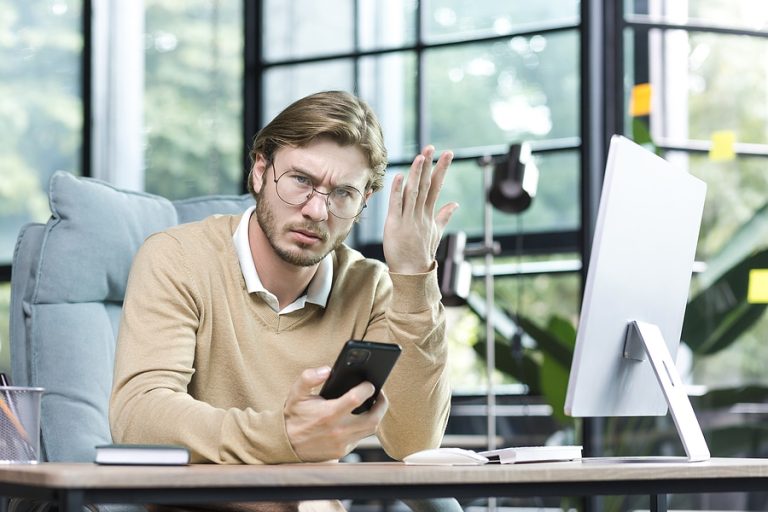 Dissatisfied businessman freelancer in glasses upset looking at camera man working inside office building holding phone and looking at camera with complaint