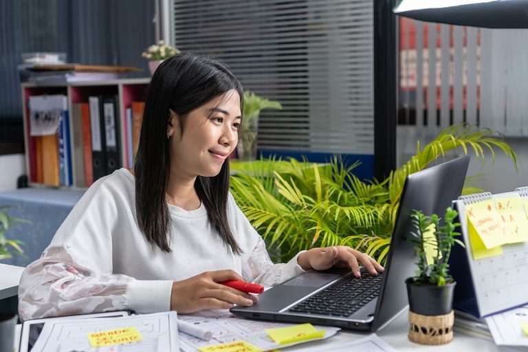 Happy Asian Young Businesswoman or University Student reading documents reports papers, entrepreneur working on line typing laptop at office computer room,smile writing notes in paperwork at desk busy