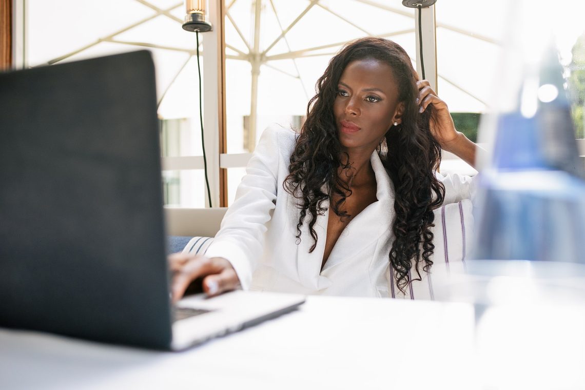 Elegant smiling African America businesswoman in white jacket browsing laptop white working remotely in cafe