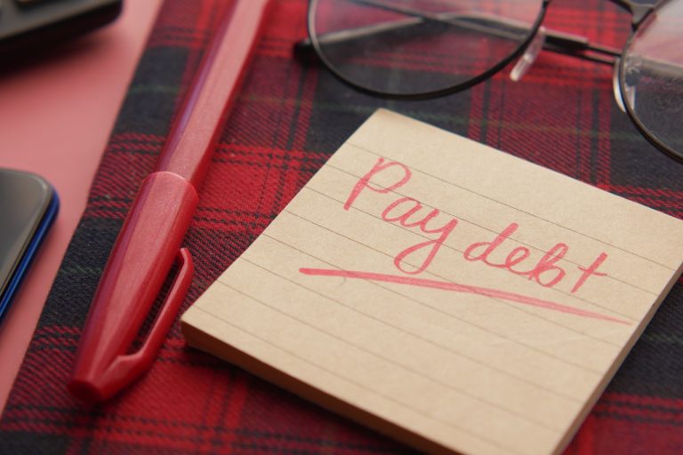 A note with "Pay debt" scribbled on it rests on a red plaid surface, accompanied by a red pen and glasses, evoking the meticulous strategy of a financial planner determined to navigate credit challenges.