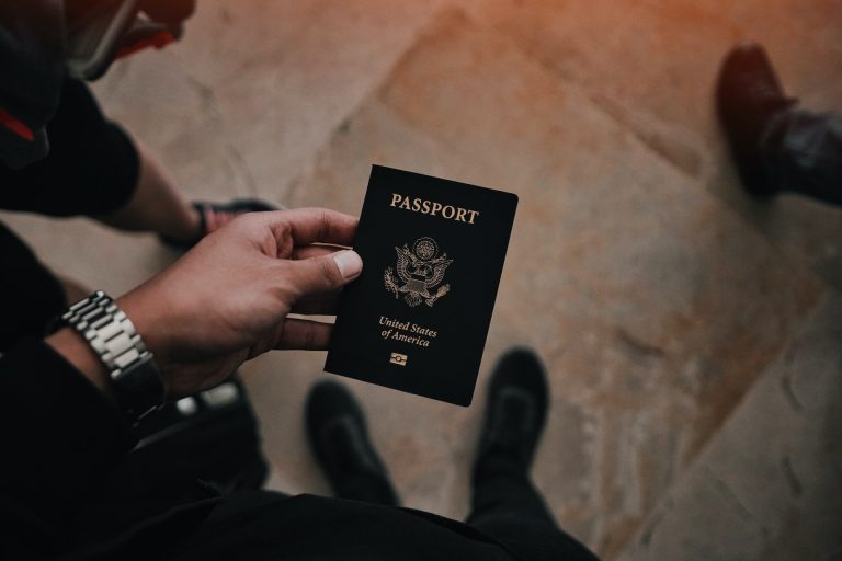 A person clutching a United States passport, wearing a watch and dark clothes, stands alongside two others, perhaps discussing budget-friendly travel tips.