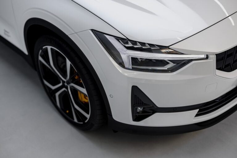 Close-up of the front left side of a white modern car with sleek lines, perfect for the budget-conscious driver. Featuring a detailed headlight and large alloy wheel on a light-colored floor, it combines style with practicality seen by any financial planner.