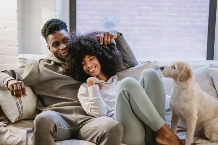 A couple sits smiling on a couch with a dog, enjoying a peaceful moment. The man, perhaps discussing their budget, has his arm around the woman, who is leaning against him. Their faithful dog looks in their direction, seemingly content with their financial planner's advice.