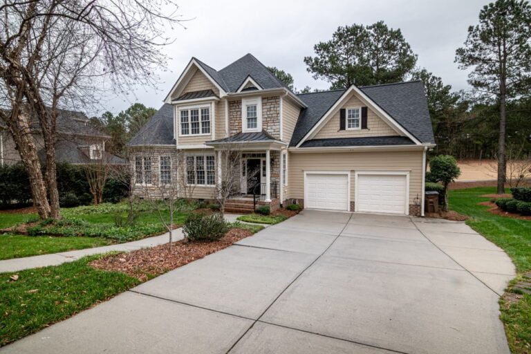 This two-story house, with gray siding and white trim under a dark shingle roof, offers both style and practicality. The spacious two-car garage is perfect for a budget-conscious family, surrounded by a manicured lawn and trees.