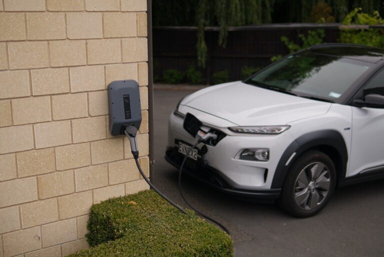 An electric car, symbolizing a budget-friendly choice, is plugged into a wall-mounted charger outside a brick building.