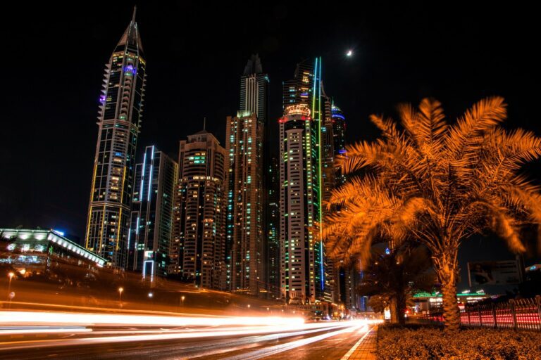 Tall skyscrapers illuminated at night with a blurred effect from moving lights below, and a palm tree in the foreground, like the bustling dreams of a financial planner carefully managing credit under the city's glowing canopy.