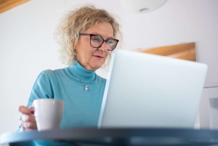 A woman with curly hair and glasses, a financial planner by profession, sips thoughtfully from her mug as she works on a laptop in a bright room.