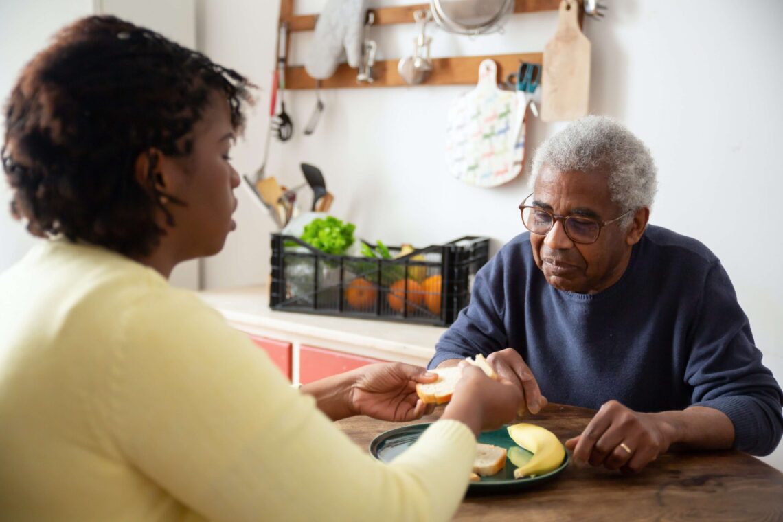 A person in a yellow sweater, perhaps a savvy financial planner, hands a piece of food to an older person seated across the table.