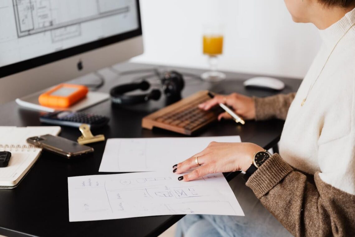 A financial planner reviews design sketches at a desk, surrounded by a computer, tablet, smartphone, and an orange drink.