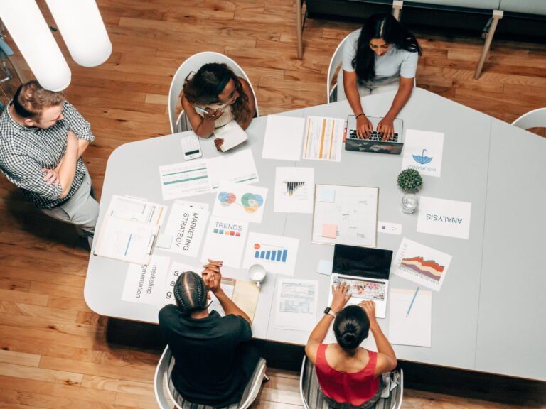 Five people gather around a table, laptops and documents in hand, discussing data and charts. Strategies unfold as they explore how to maximize your marketing budget efficiently.