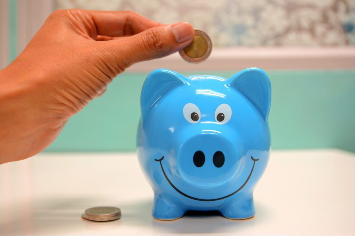 A hand is carefully placing a coin into a blue piggy bank, with two coins beside it on the white surface, illustrating a smart approach to budgeting.