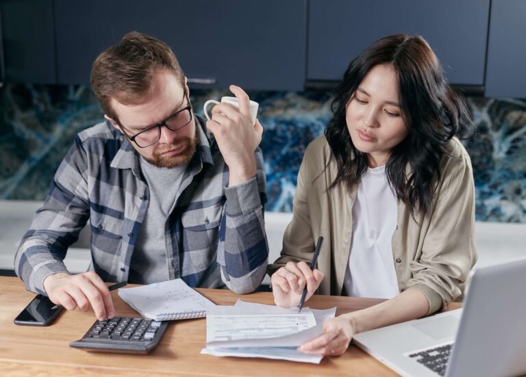 A man and woman sit at a table, meticulously reviewing documents. Acting as a financial planner, the man holds a mug while using a calculator, as the woman writes on the papers. A laptop and smartphone lie nearby, hinting they are analyzing their budget.