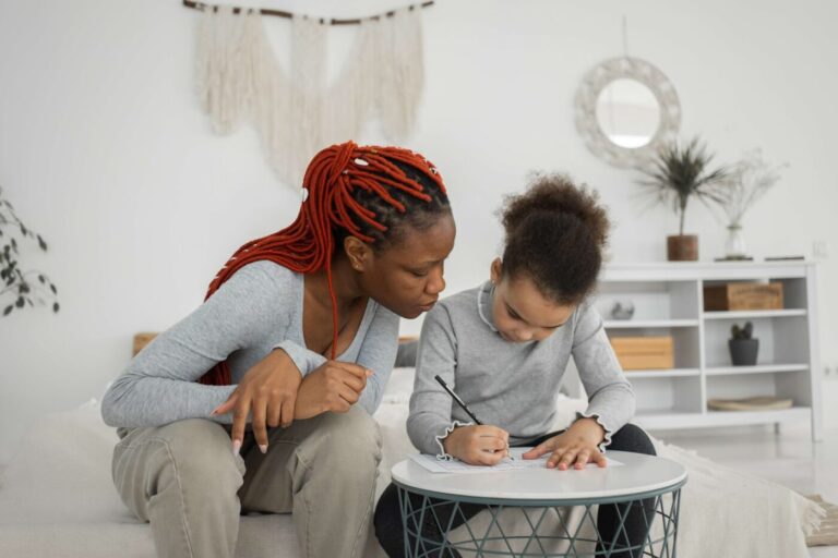 An adult assists a child with drawing at a round table in a cozy, well-decorated room, much like how a financial planner guides clients through their budget.