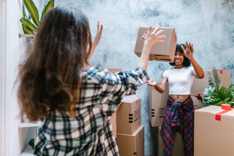 Two people in a room with stacked cardboard boxes; one person is tossing a box to the other, who is wearing a white shirt and plaid pants. A plant is visible in the background, as they work together on organizing within their budget.