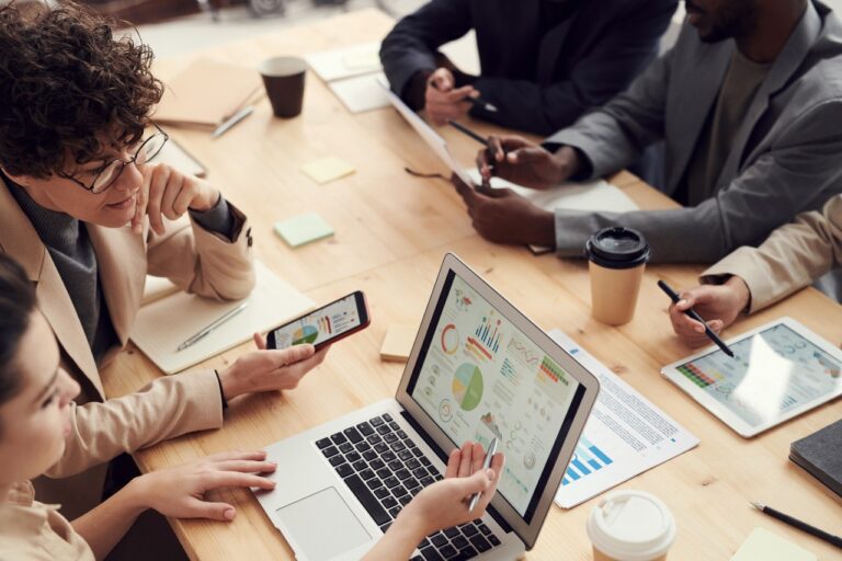 A group of people at a wooden table engaged in a discussion, with laptops, papers, and coffee cups visible. A financial planner's laptop screen displays colorful charts and graphs, likely aiding in budget discussions.