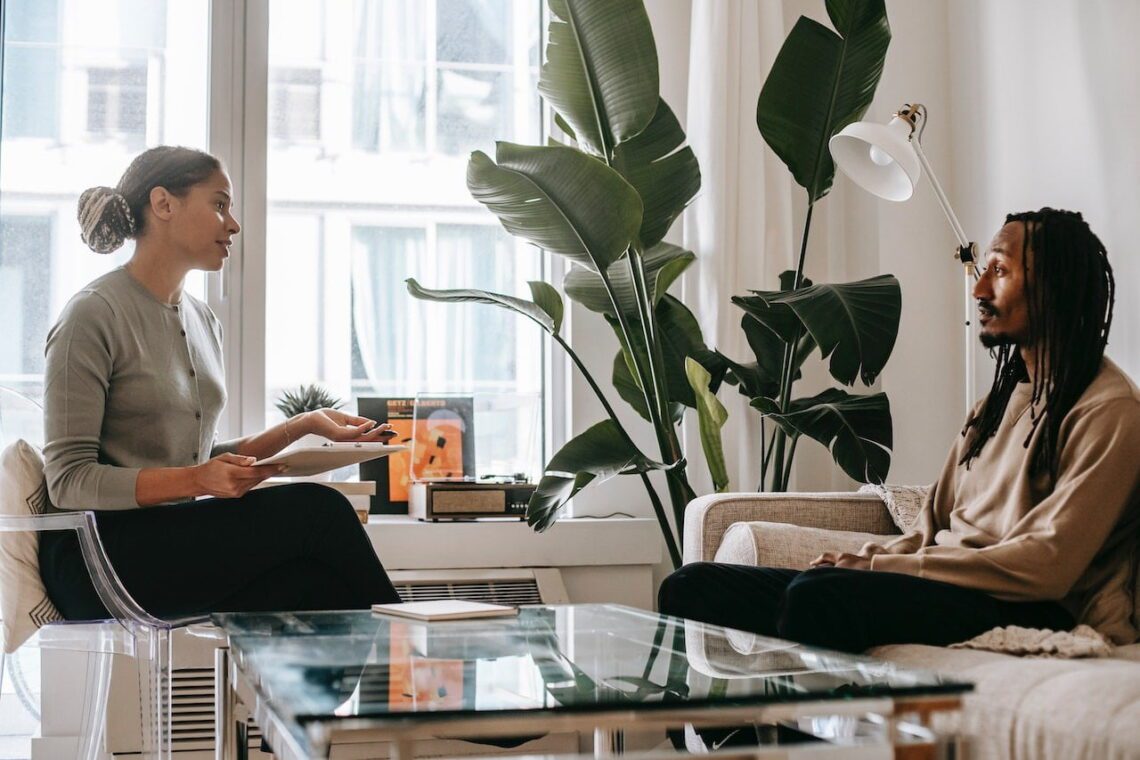 Two people sit in a living room with large plants, discussing their budget in a relaxed setting. One holds a tablet, perhaps reviewing their financial planner's advice to keep their credit in check.