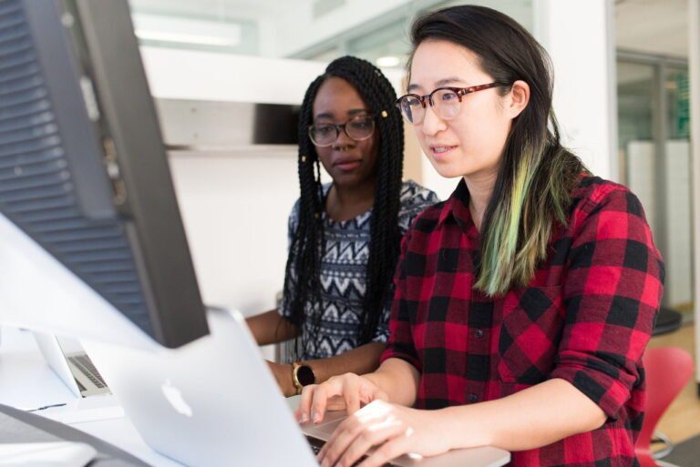 Two people work at computers in an office. One, a financial planner in a red plaid shirt, focuses on spreadsheets, while the other, wearing glasses and a patterned shirt, drafts credit reports.