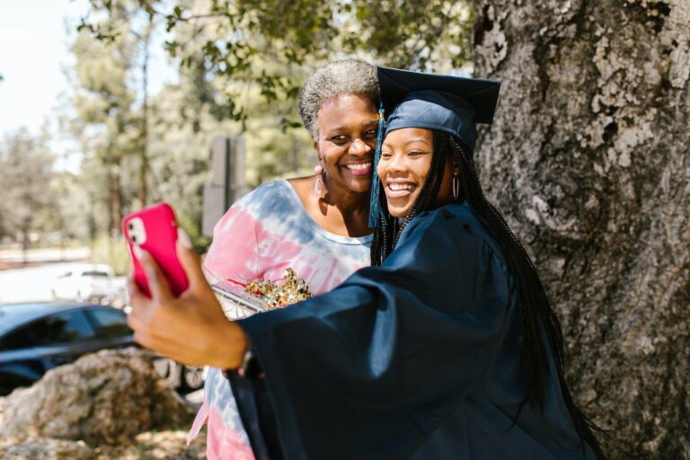 A graduate in cap and gown snaps a selfie with a financial planner under the shade of a large tree, celebrating not just academic success but also sound budgeting for the future.