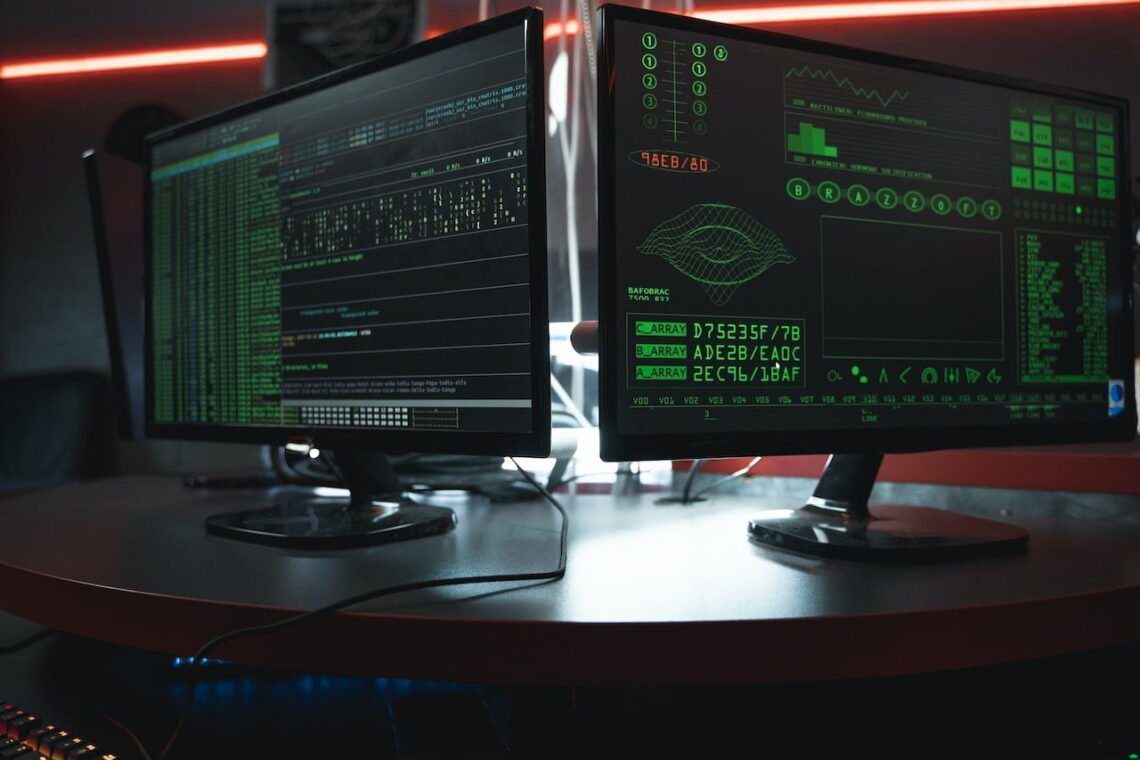 Two computer monitors displaying green text and graphics, showing financial planner data and system analysis, on a desk in a dimly lit room with red lighting.