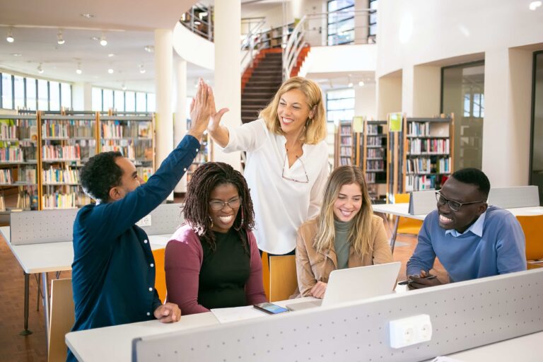 A group of five people in a library smiles and high-fives at a table with a laptop, surrounded by bookshelves, celebrating their new budget plan.