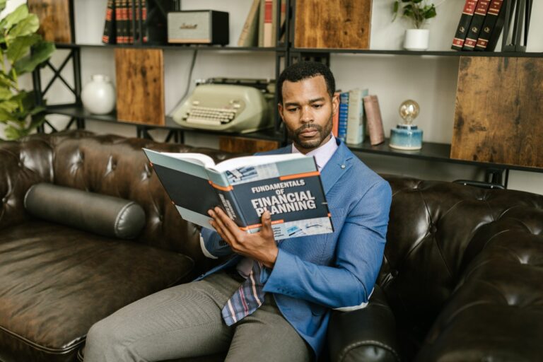 A financial planner in a blue blazer sits on a brown leather couch, immersed in "Fundamentals of Financial Planning," surrounded by bookshelves and a vintage typewriter, contemplating credit strategies.